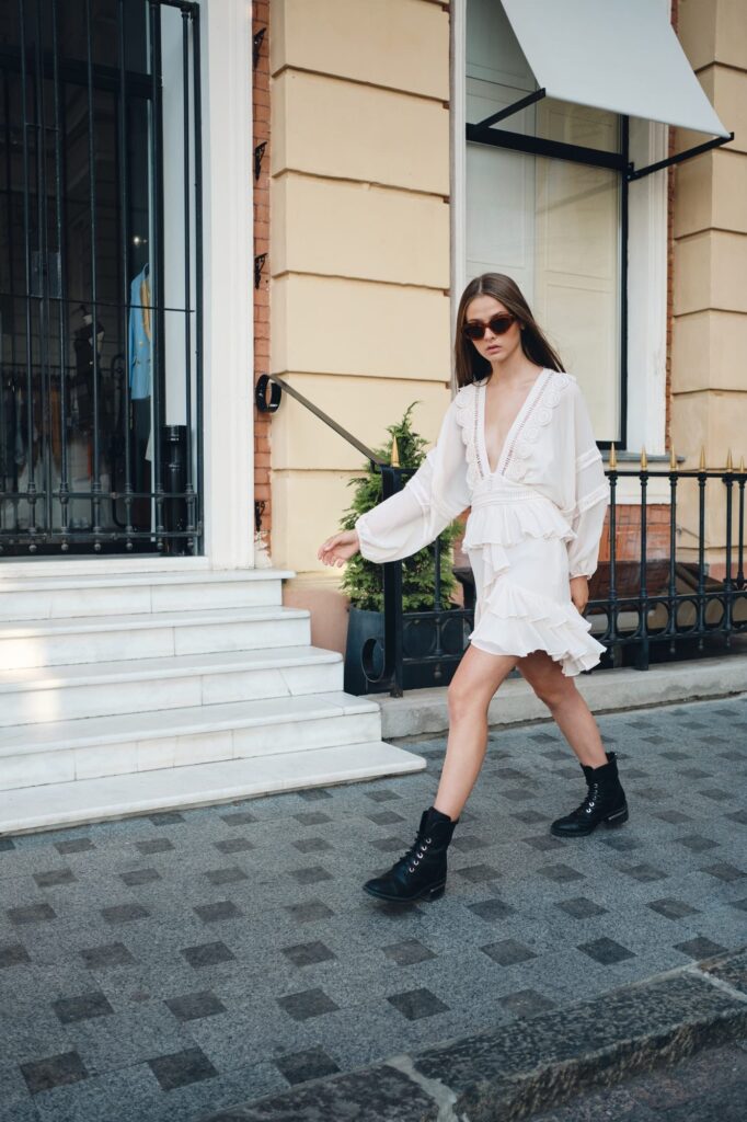 Young model in sunglasses, white dress and boots thoughtfully looking in camera walking on street